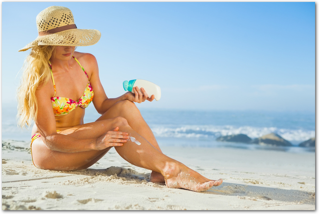 Gorgeous woman sitting on the beach in sunhat applying suncream on a sunny day
